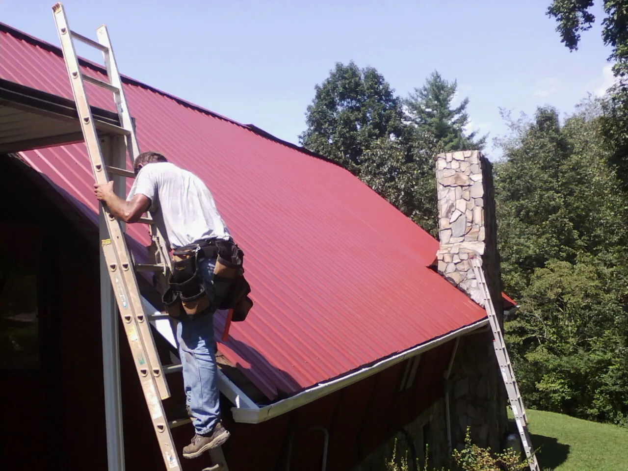 Roofer on a ladder beside a red metal roof and stone chimney — Soffit & Fascia Repair by Roofing Brothers in Topsfield